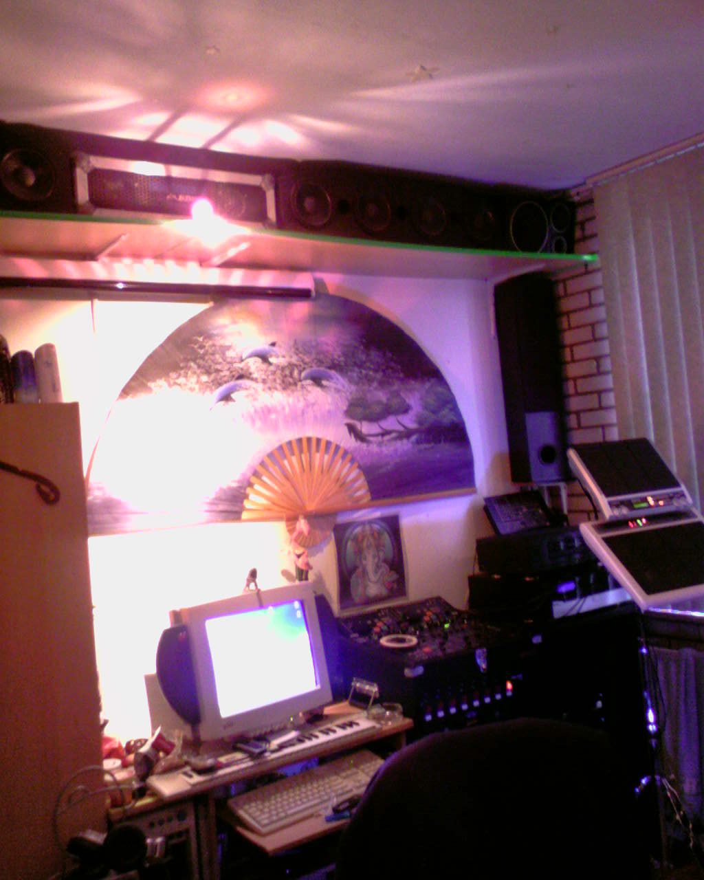 A home studio room with speakers mounted on the wall, a large CRT monitor, mixing gear and coloured light